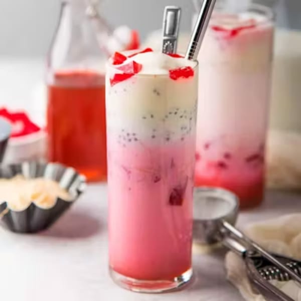 A tall glass of pink and white layered Thai dessert with red jelly and basil seeds.