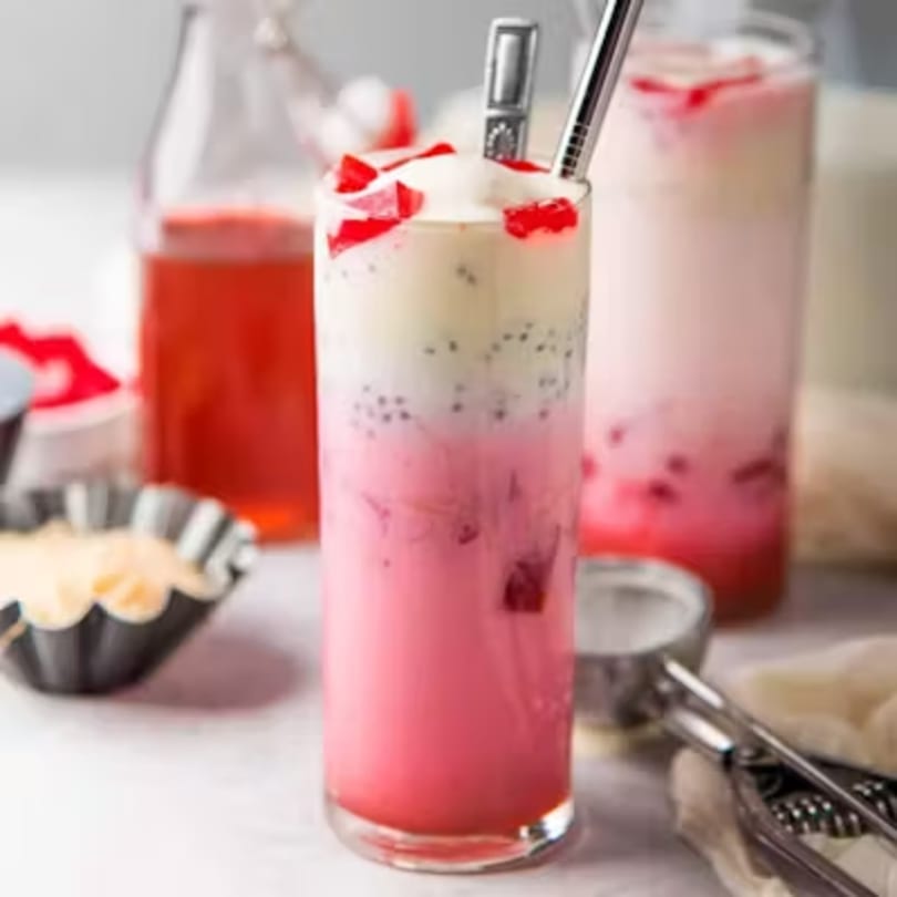 A tall glass of pink and white layered Thai dessert with red jelly and basil seeds.