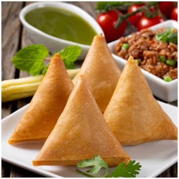 Four golden-brown samosas on a white plate, with green sauce and minced meat in the background.