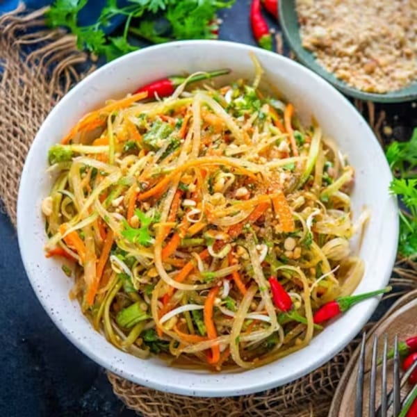 A vibrant Thai papaya salad with shredded vegetables, peanuts, and red chilies in a white bowl.