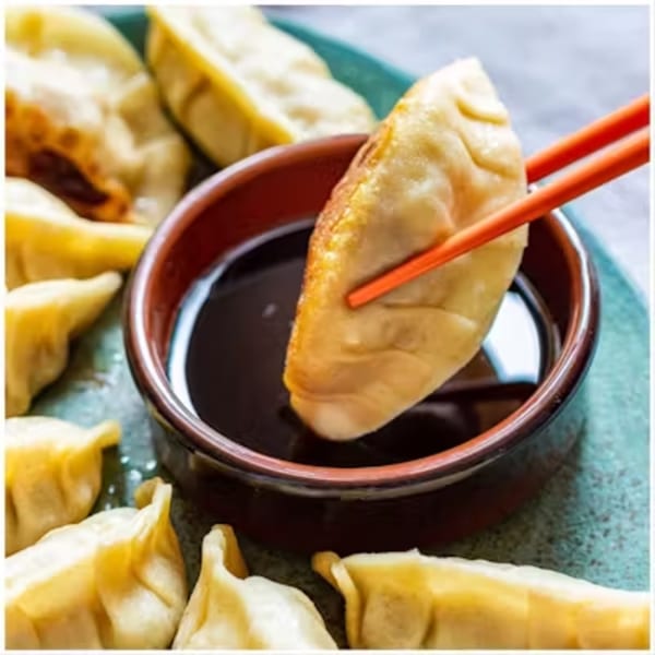 Chopsticks dipping a golden-brown dumpling into a small bowl of dark sauce, with more dumplings nearby.